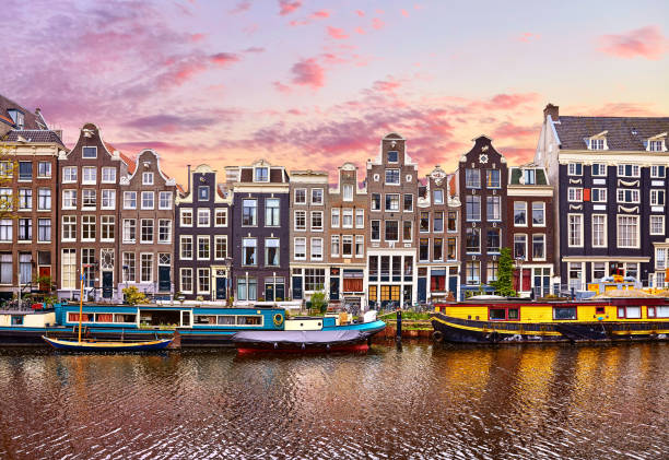 Amsterdam Amsterdam, Netherlands. Floating Houses and houseboats and boats at channels by banks. Traditional dutch dancing houses among trees. Evening autumn street above water pink sunset sky with clouds.